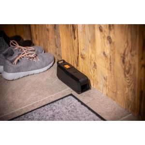 Windhager elektronische Mausefalle, schwarzes Gehäuse mit orangem Knopf, neben Schuhen an einer Wand platziert.