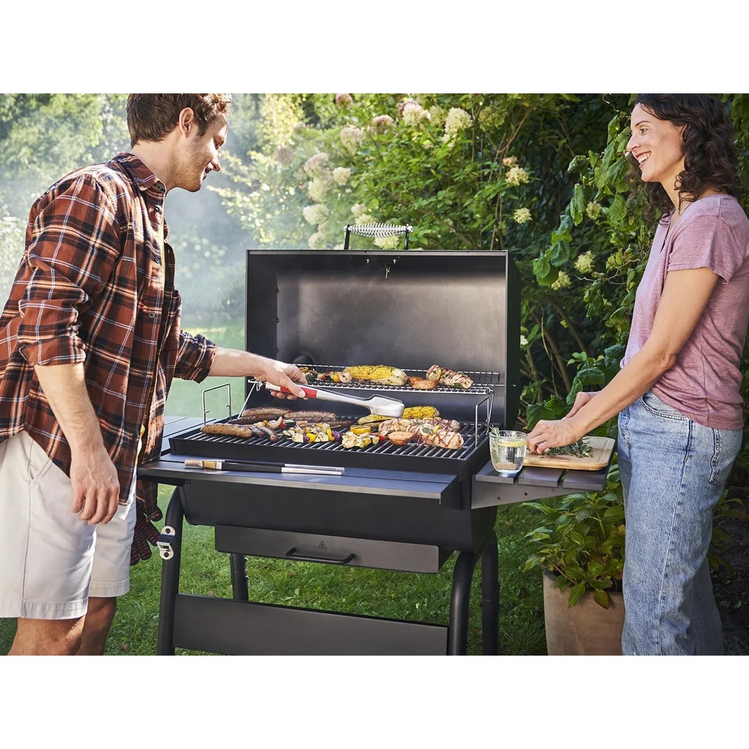 Char-Broil Holzkohlegrill Charcoal L beim Grillen von Würstchen, Mais und Gemüse im Garten.