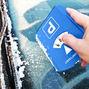 Hand kratzt mit blauer Parkscheibe und integriertem Eiskratzer Eis von vereister Autoscheibe. Praktischer Frostschutz.