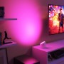 Weiße Philips Smart-LED-Tischleuchte Squire Lite projiziert pinkes Wallwashing-Licht auf eine Wand neben einem TV. Schafft Ambiente.