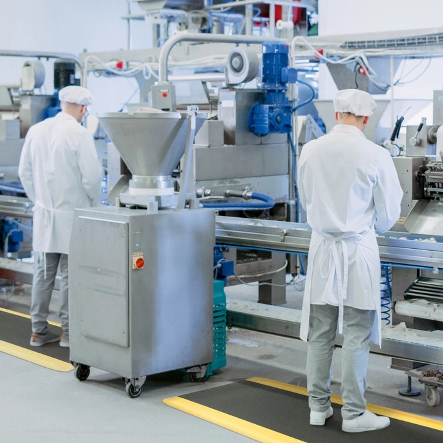 Schwarze KARAT Anti-Ermüdungsmatte mit gelbem Rand in Produktionshalle, zwei Arbeiter im Hintergrund.