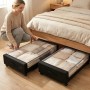 Frau zieht schwarze Unterbettkommode mit transparentem Deckel und Rollen unter Bett hervor, gefüllt mit Kleidung.