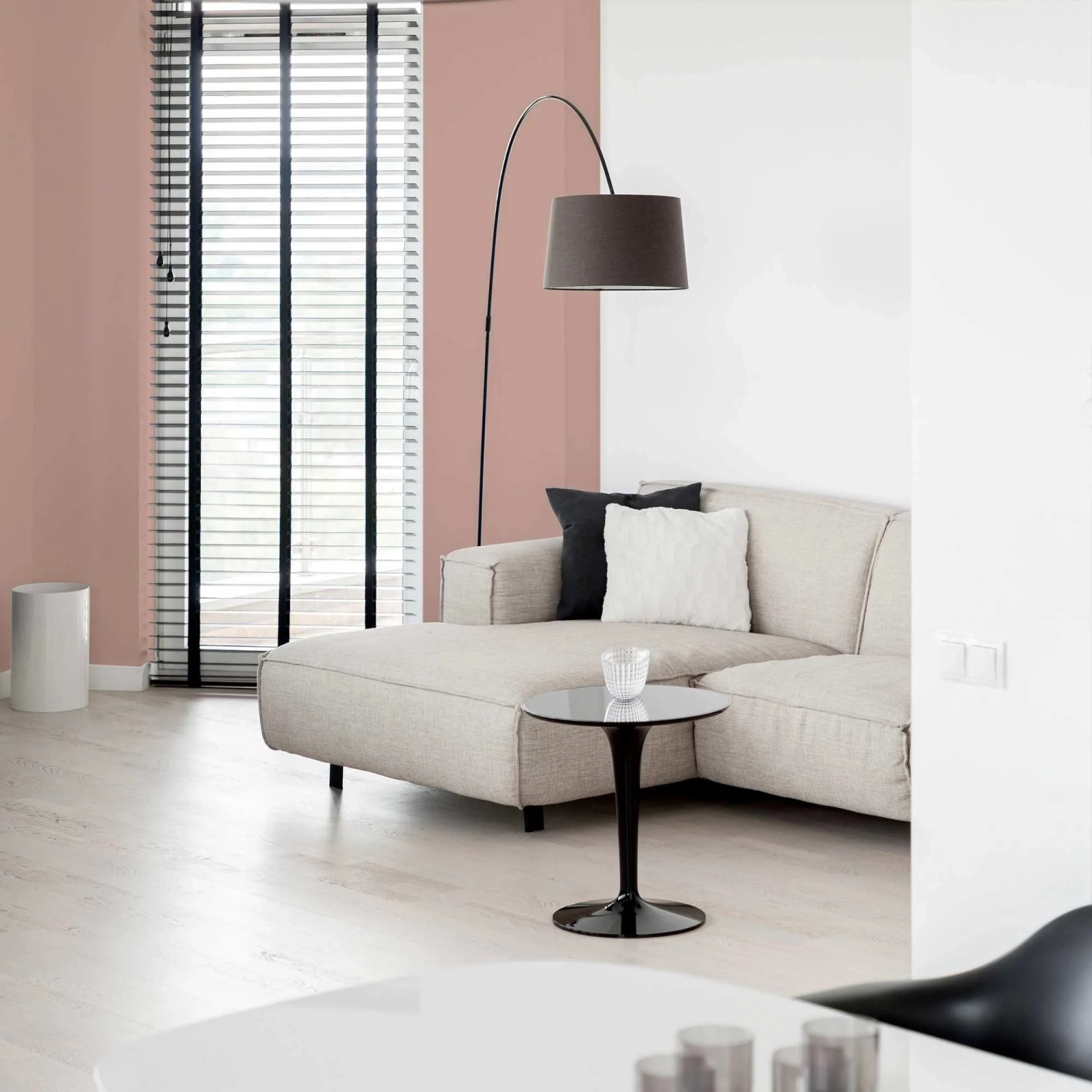Tadessi Wandfarbe Taupe matt in modernem Wohnzimmer. Eine Wand ist in warmem Taupe gestrichen, mit Sofa, Lampe und Fenster.