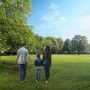 Familie mit Picknickkorb geht durch einen Park mit grüner Wiese und Bäumen.