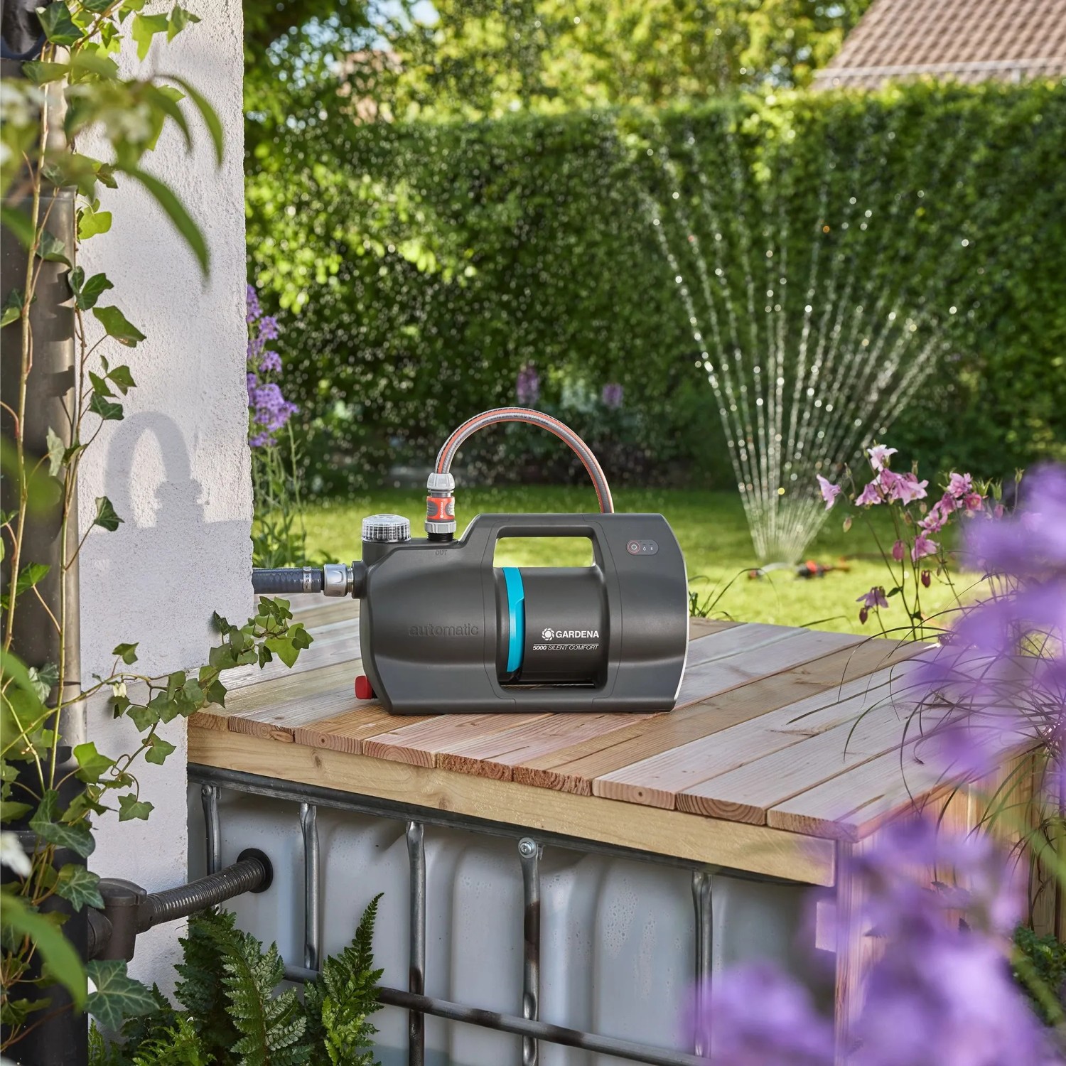 Gardena Haus- und Gartenautomat 5000 Silent Comfort auf Holzpodest, Rasensprenger im Hintergrund.