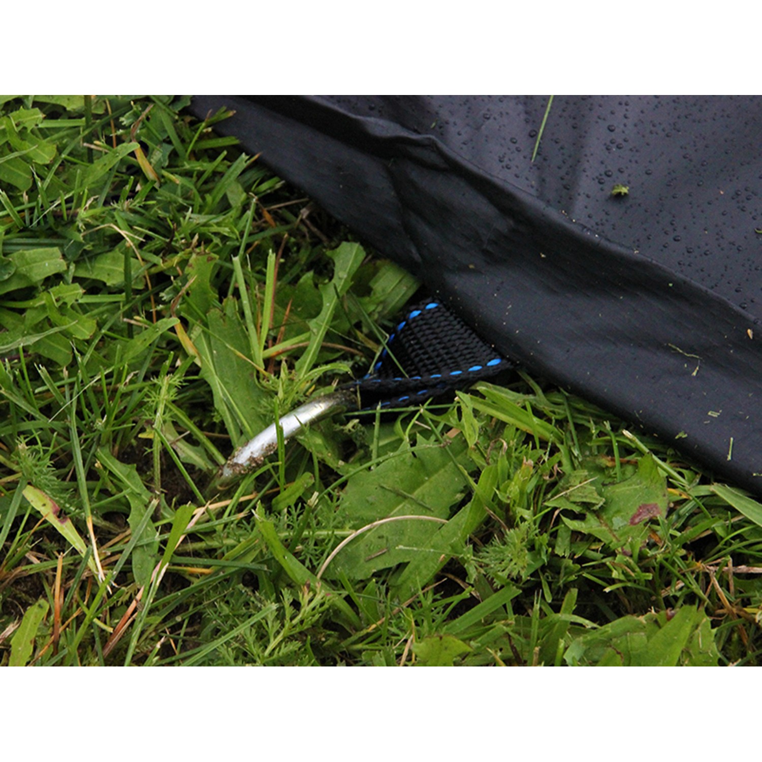 Schwarze Abdeckplane für rechteckiges EXIT Trampolin, befestigt mit Hering im Gras.