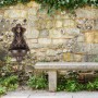 Antiker Gartenbrunnen aus bronzefarbenem Aluminiumguss mit Goldakzenten und Wasserhahn an Steinmauer.