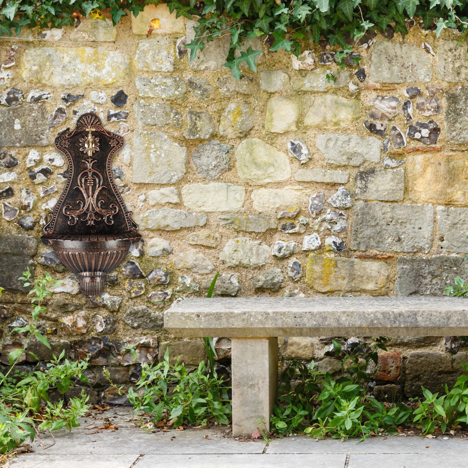 Antiker Gartenbrunnen aus bronzefarbenem Aluminiumguss mit Goldakzenten und Wasserhahn an Steinmauer.