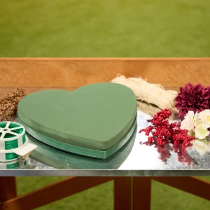 Grünes herzförmiges Steckschaumherz für frische Blumen, umgeben von Bastelmaterialien. Ideal für kreative Blumengestecke.