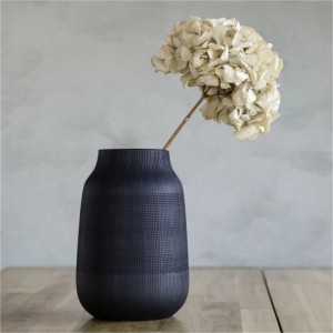 Dunkle, strukturierte Vase mit getrockneter, cremefarbener Blume auf Holztisch vor gemusterter Wand.