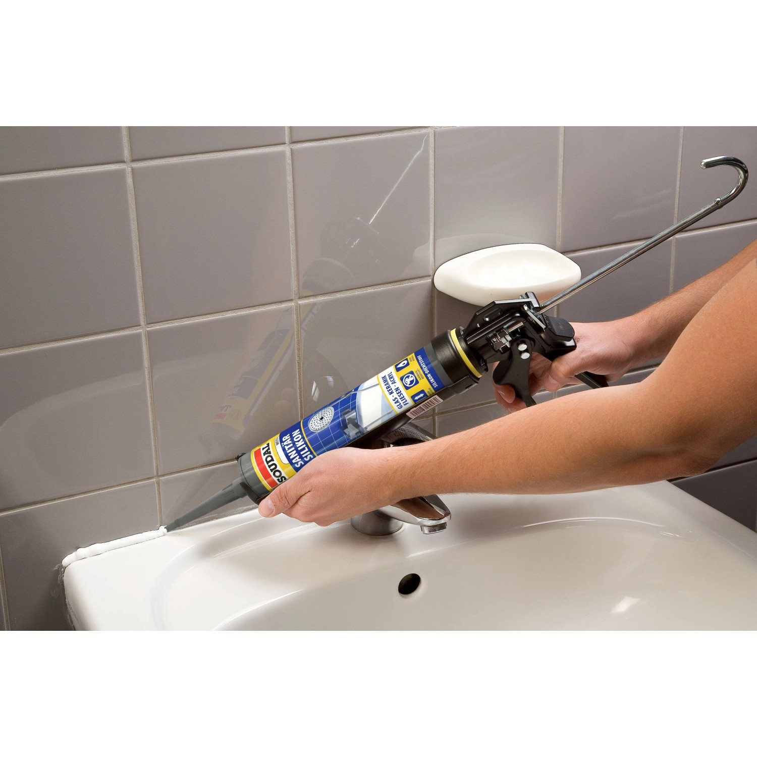 Person dichtet Waschbecken mit weißem Soudal Sanitär Express Silikon aus Kartuschenpistole ab. Fliesenwand im Bad.