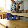 Mann bohrt in Holzbrett, das von blauem Wiltec Parallelschraubstock 125mm auf Werkbank gehalten wird.