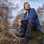 Mann sitzt mit Rucksack am Flussufer, Tesa Reparaturband Ultra Power Extreme für Outdoor-Reparaturen.