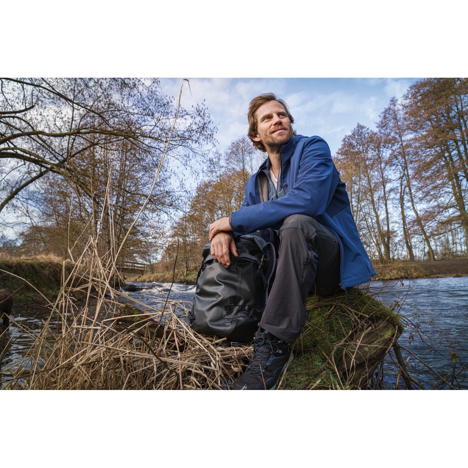 Mann sitzt mit Rucksack am Flussufer, Tesa Reparaturband Ultra Power Extreme für Outdoor-Reparaturen.