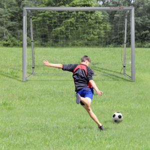 Graues SPORTNOW Fußballtor aus Stahl mit Netz auf Rasen. Ein Spieler schießt Ball aufs Tor. Ideal für Sport & Training.