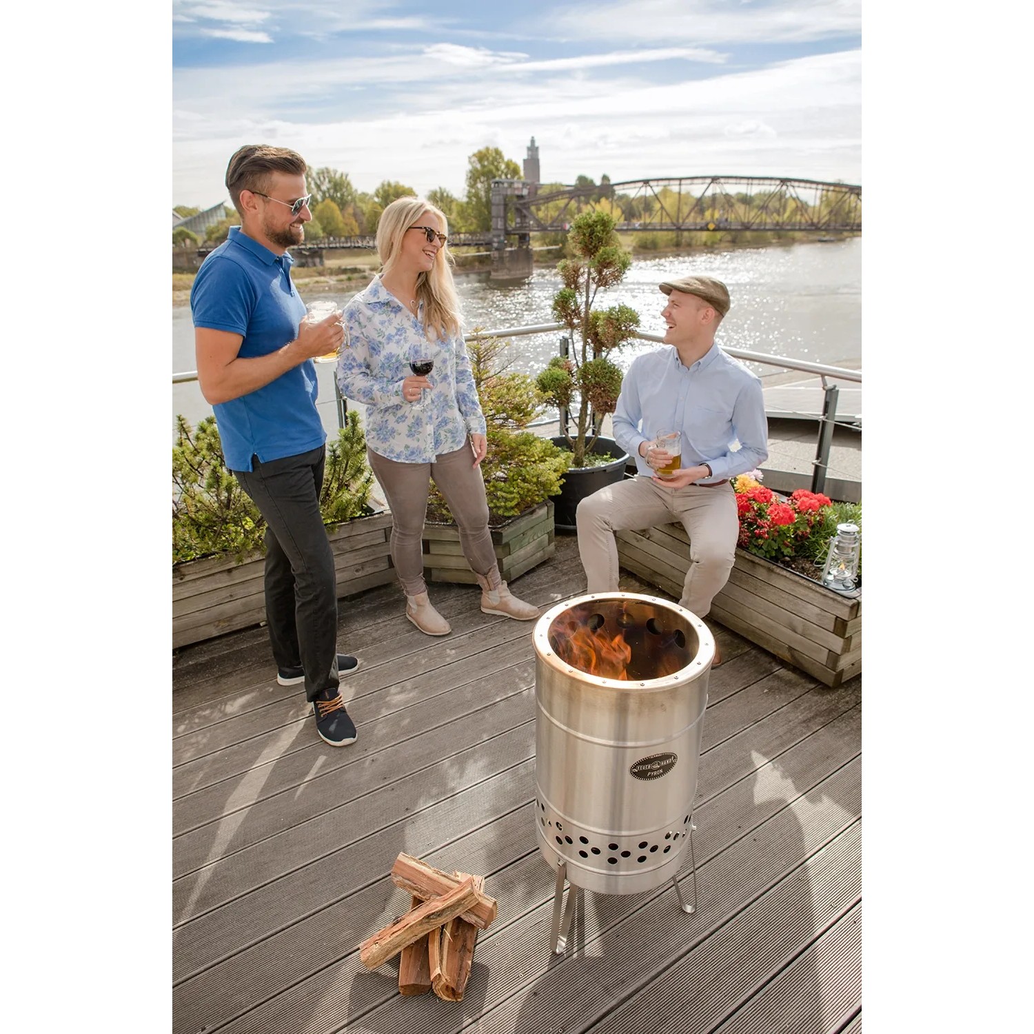 Feuerhand Pyron Feuertonne aus Edelstahl (25 l) mit drei Personen im Hintergrund.