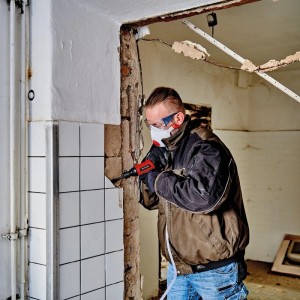 Mann arbeitet mit Einhell Kompressor an Wandfliesen, Schutzbrille und Maske. Abbruchhammer im Einsatz.