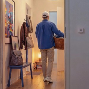Weißer PHI Bewegungssensor an Flurwand, aktiviert Licht für Person mit Wäschekorb. Smarte Leuchtmittel.