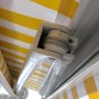 Detailaufnahme: Sandfarbene MCW Kassettenmarkise mit gelb-weißen Streifen, Seilzug und Mechanik.