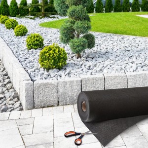 Schwarzes KARAT Unkrautvlies Ground Control Rolle mit Schere vor Gartenbeet mit Steinen.