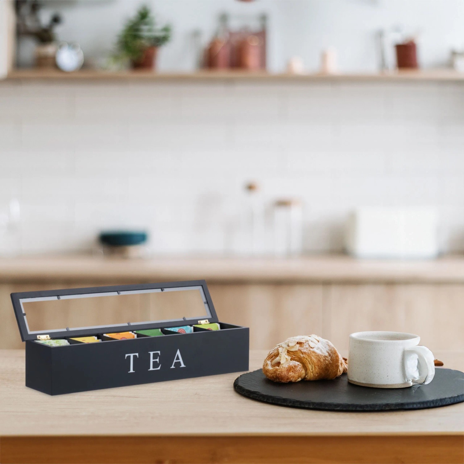 Schwarze Bambus Teebox mit 6 Fächern, transparentem Deckel und „TEA“-Aufschrift auf Küchentheke, gefüllt mit Teebeuteln.