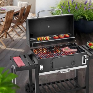 Schwarzer Taino Hero XXL Holzkohlegrill mit offenem Deckel, Grillgut, Seitentischen und Rollen auf Terrasse.
