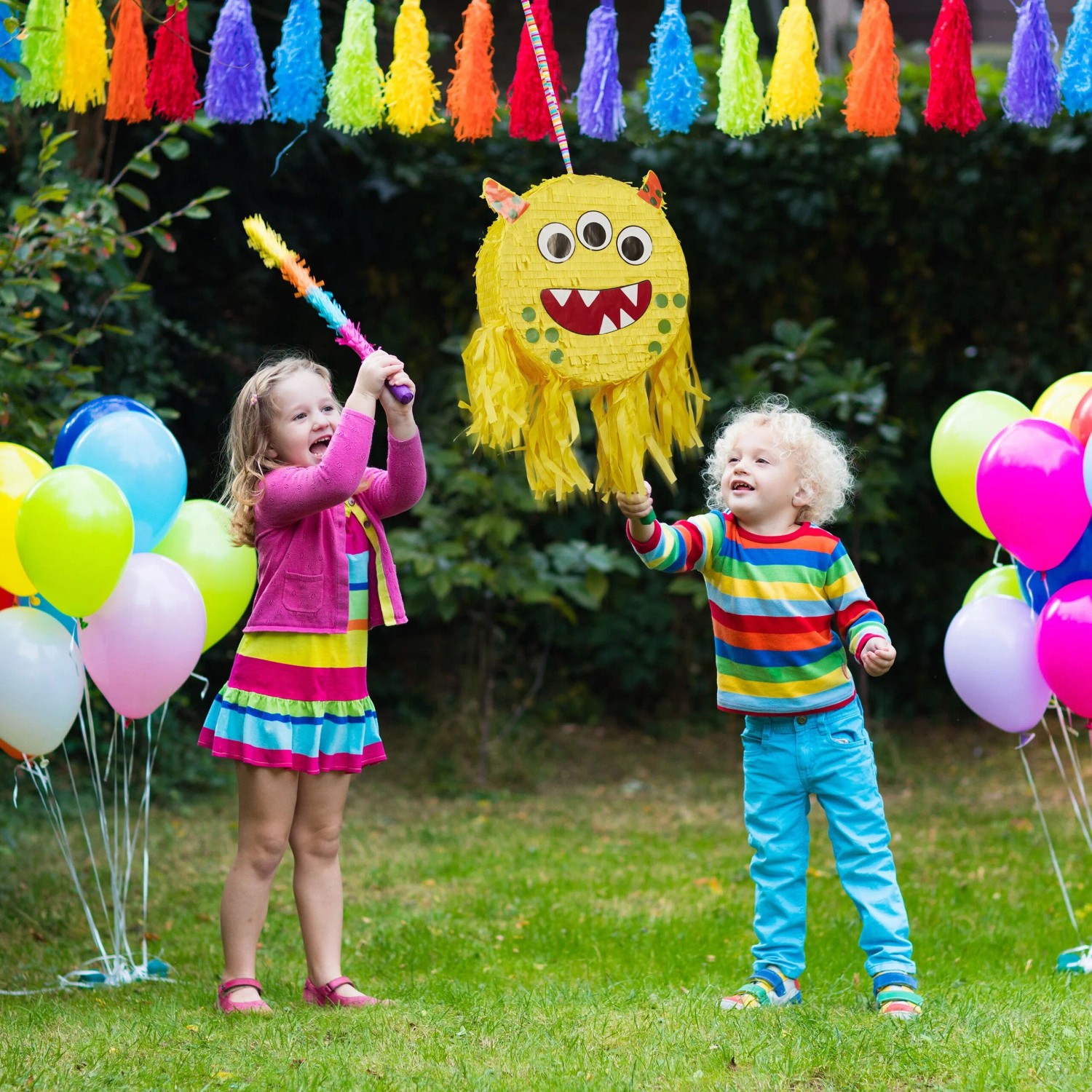 Zwei Kinder spielen im Garten mit einer gelben Monster Pinata aus Pappe. Bunte Partydekoration für Kindergeburtstag.