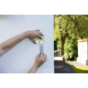 Außenaufnahme: Person befestigt mit Uhu Repair All Powerkit die Hausnummer 1 an einer weißen Wand.