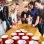 Fröhliche Freunde spielen Bierpong mit roten Kunststoffbechern und Bällen auf einem Holztisch. Das Bierpong Set für Partys.