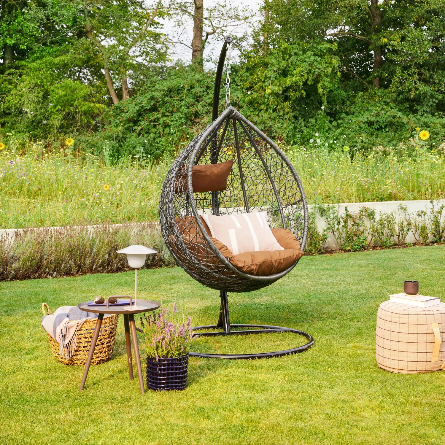 Grauer Home Deluxe Hängesessel Cielo aus Polyrattan mit Gestell und Kissen im Garten.