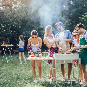 Freunde grillen im Garten, im Hintergrund ein weißer Klapp-Barhocker und Stehtisch für Outdoor-Feiern.