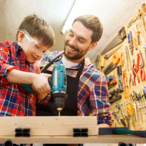 Mann und Kind bohren Holz an einer klappbaren Werkbank mit Spannbacken in der Werkstatt. Kind trägt Schutzbrille.