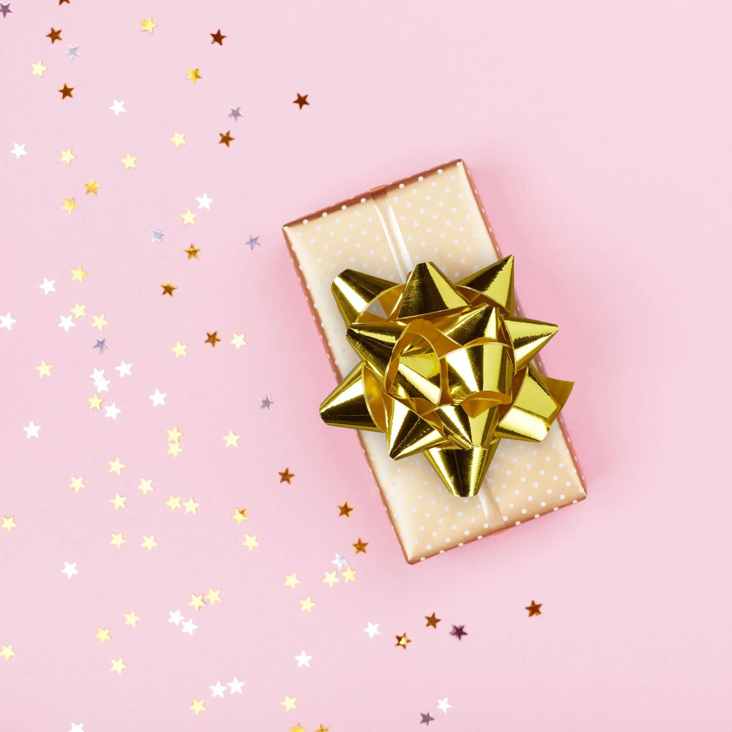 Glänzende goldene Geschenkschleife auf beigem Geschenk mit Punkten, umgeben von Sternenkonfetti. Perfekt für Weihnachtsdekoration.