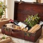 Geöffnete Holztruhe im Vintage-Stil mit Wein, Gebäck und Blumen als Geschenkidee.