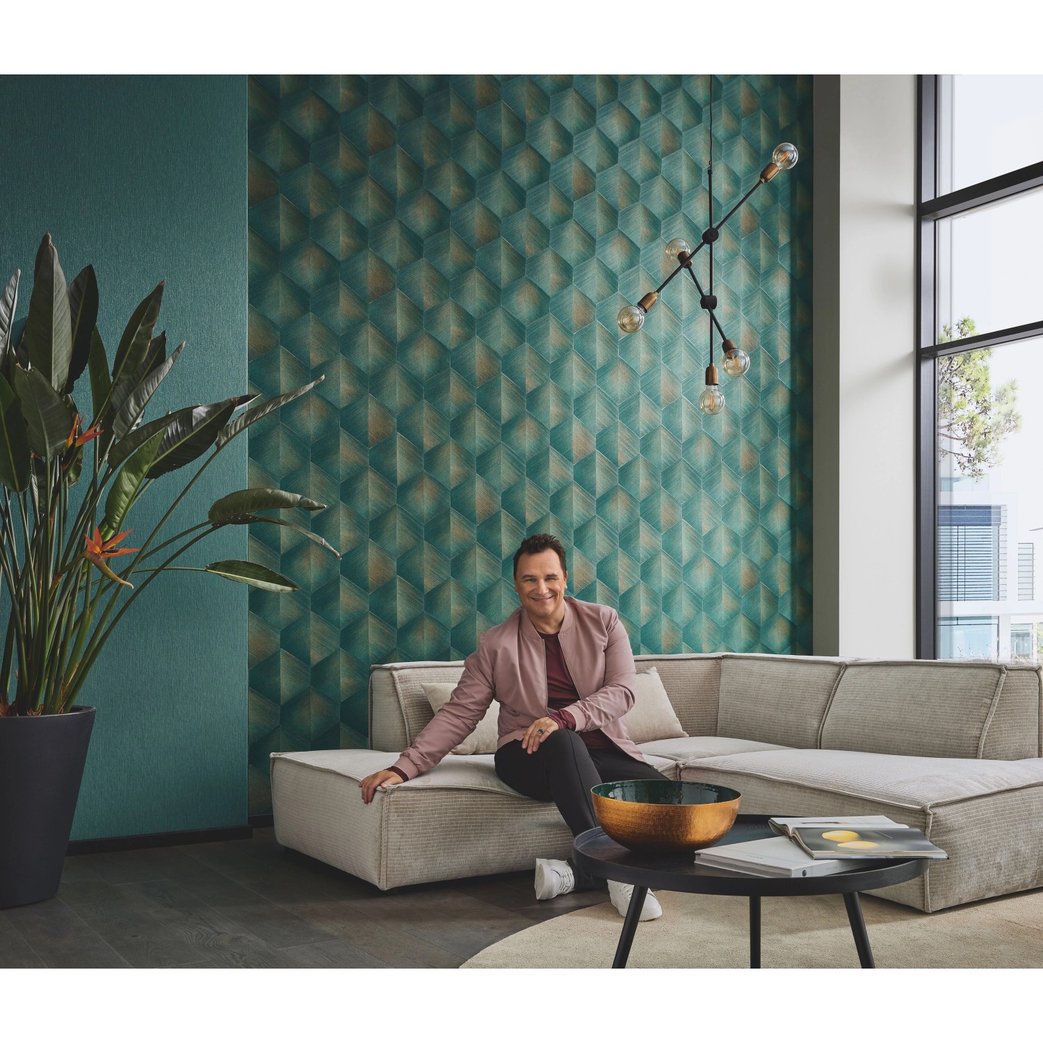 Guido Maria Kretschmer Vliestapete Prisma Petrol im Wohnraum mit Person auf Sofa.