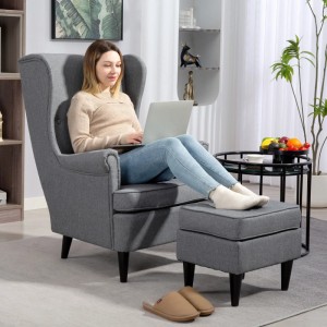 Grauer Ohrensessel mit Hocker im Wohnzimmer, Bezug in Leinenoptik. Frau sitzt mit Laptop im Sessel.