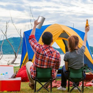 Großer silberner Edelstahl-Flachmann beim Camping. Ein Paar sitzt vor Zelt und See, Mann hält Flachmann hoch.