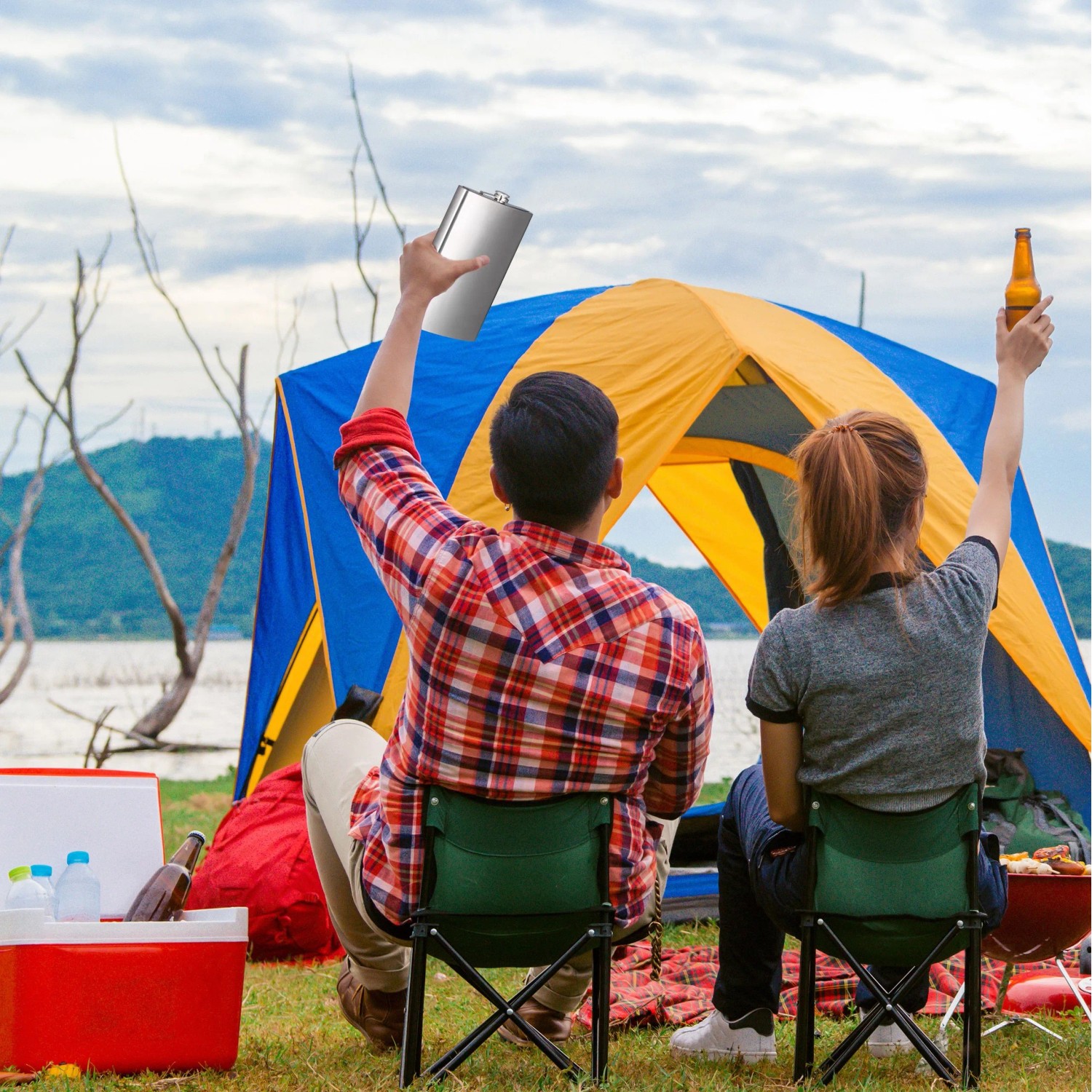 Großer silberner Edelstahl-Flachmann beim Camping. Ein Paar sitzt vor Zelt und See, Mann hält Flachmann hoch.