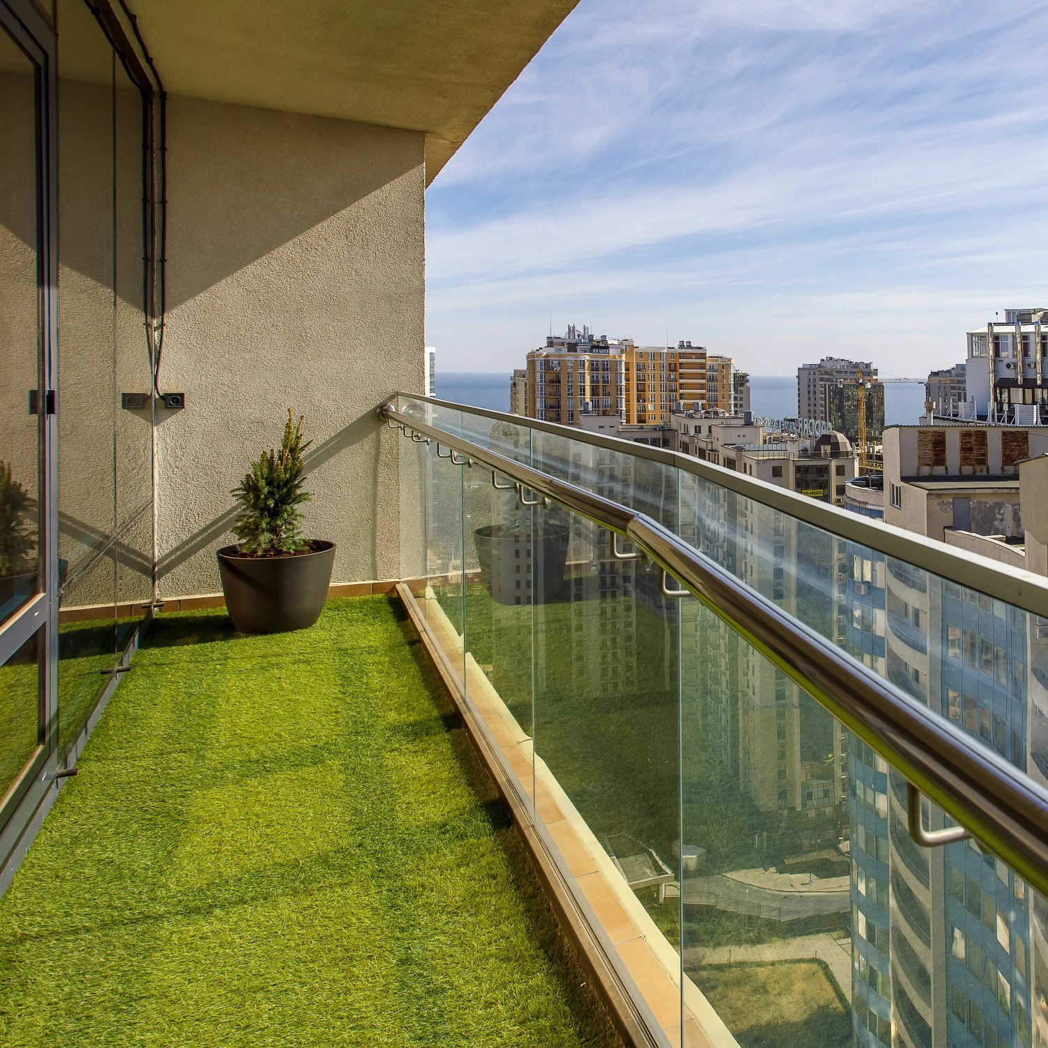 Balkon mit Primaflor Kunstrasen Cyprus: Grüner Kunstrasen mit Pflanze auf einem sonnigen Balkon.