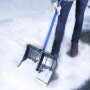 Person schiebt Schnee mit blauer Teleskop-Schneeschaufel aus Stahl und Kunststoff auf gepflastertem Weg.