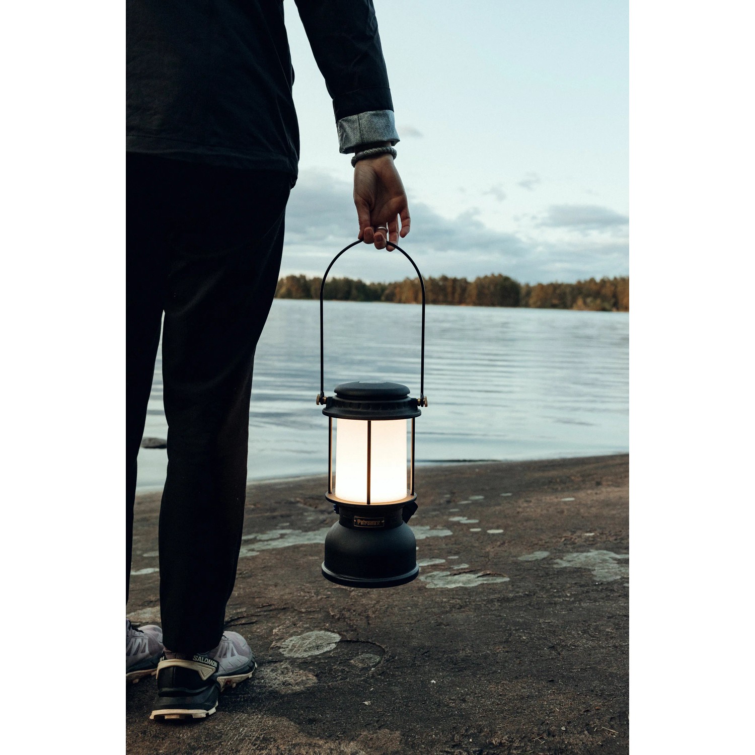 Schwarze Petromax LED-Laterne PX-LM500, gehalten von einer Person vor einem See. Camping Leuchte mit 160h Leuchtdauer.