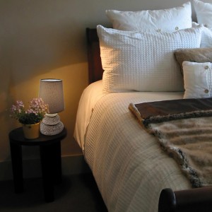 Braune Näve Tischleuchte Stoney aus Keramik, 31 cm hoch, neben einem Bett auf einem Nachttisch.