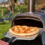 Ooni Karu 2 Pizzaofen mit Pizza auf Pizzaschieber vor Garten. Gasgrill für Holz, Kohle oder Gas.