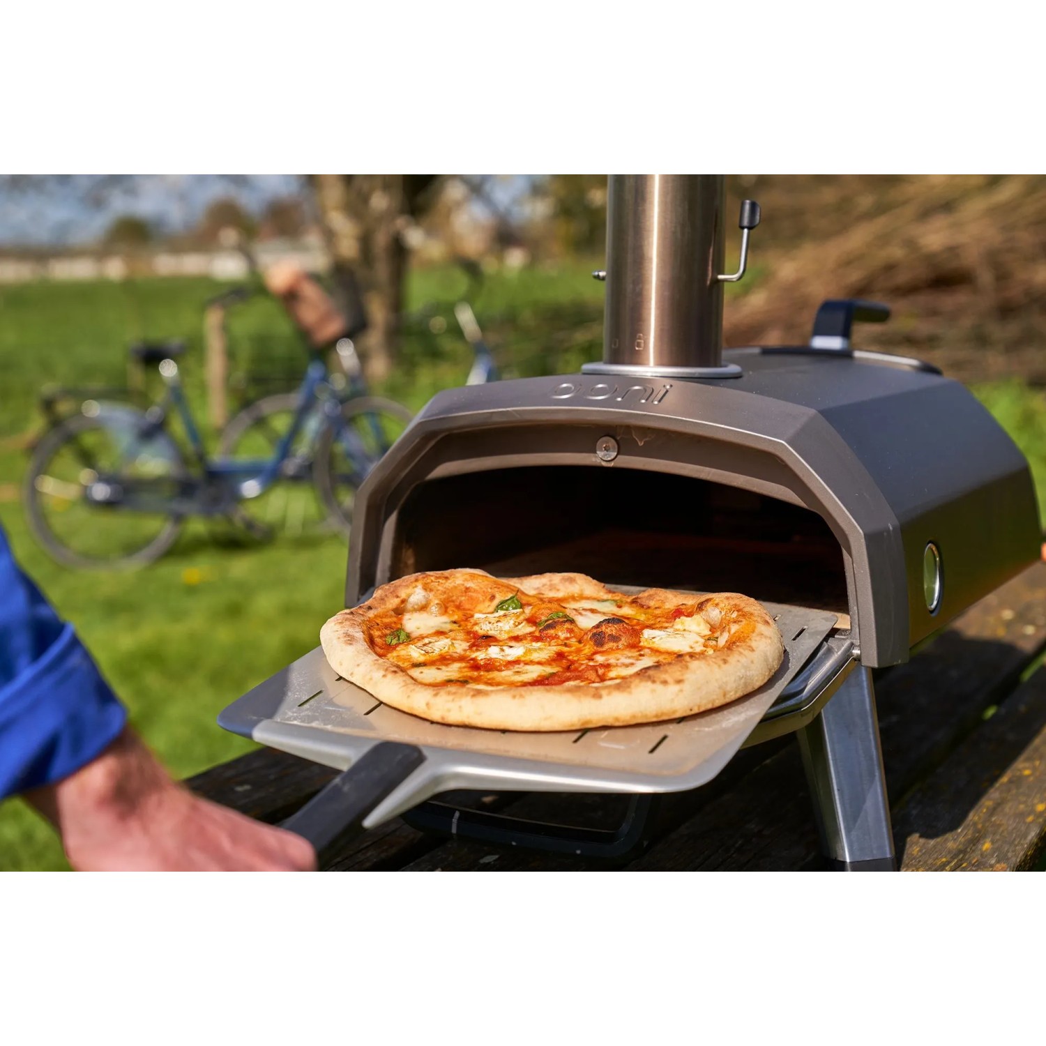 Ooni Karu 2 Pizzaofen mit Pizza auf Pizzaschieber vor Garten. Gasgrill für Holz, Kohle oder Gas.