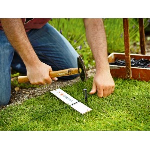 Gardena Begrenzungskabel Installation: Mann befestigt Draht für Mähroboter mit Hammer im Garten.