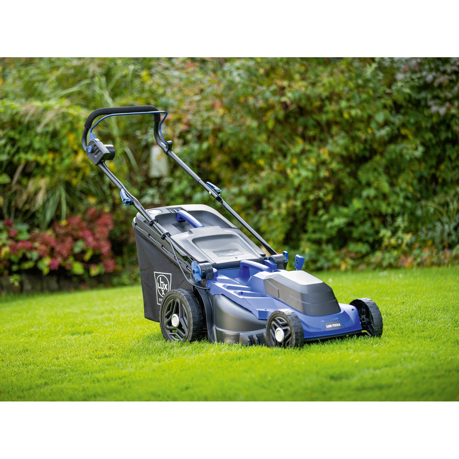 Blauer LUX-TOOLS Elektro-Rasenmäher E-RM-1800/43 auf Rasen vor Büschen.