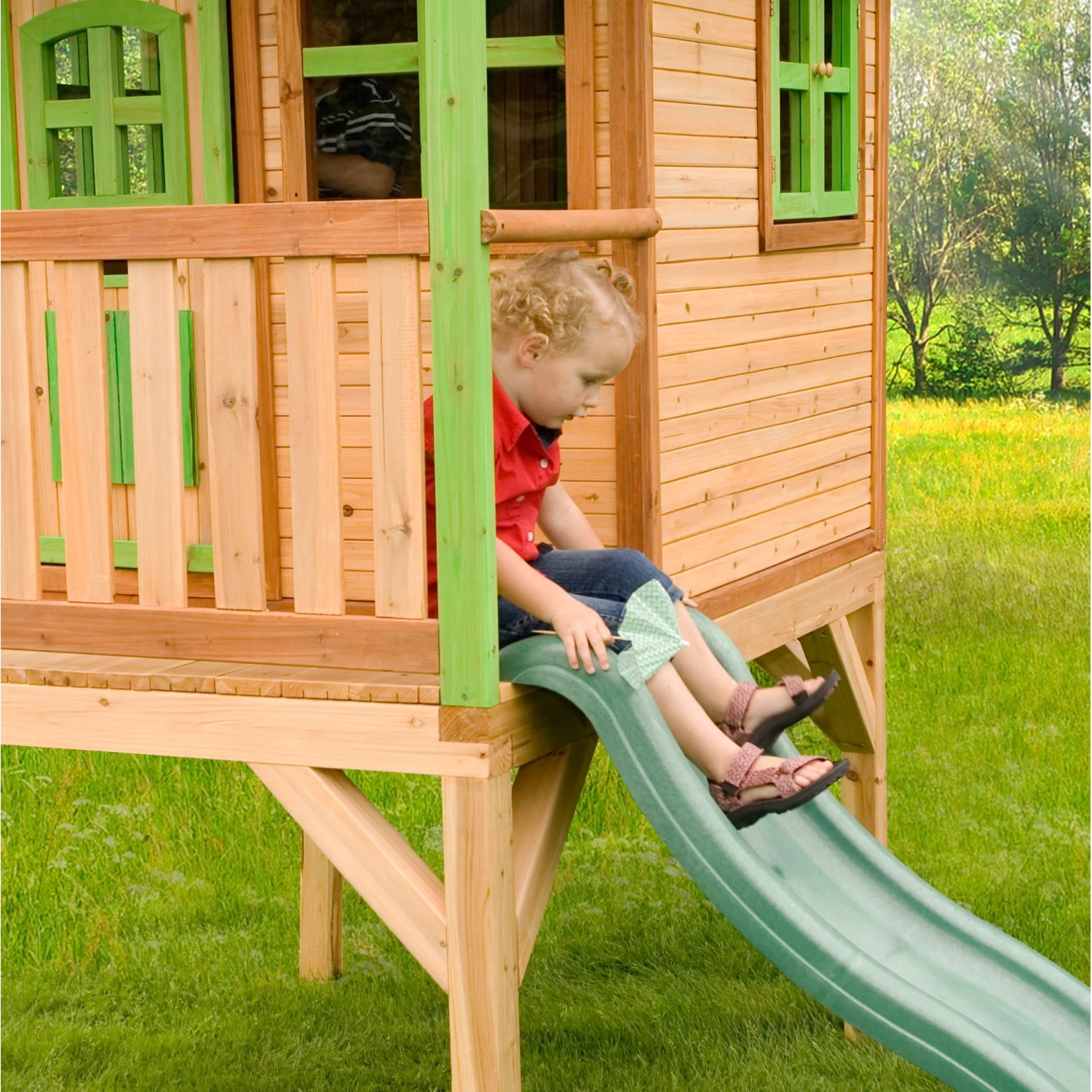 Kind auf Rutsche des Spielhauses Laura mit Leiter. Holzhaus mit grünen Akzenten im Garten.
