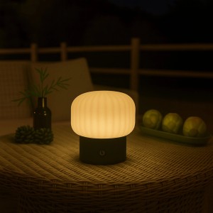 Brilliant LED Tischleuchte Sarastro, weiß-gerippt, schwarz, leuchtet warm auf einem Rattantisch im Dunkeln. Ideal für Balkon & Terrasse.