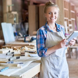 Lächelnde Frau mit Schürze und Tablet in einer Werkstatt, umgeben von Holz und Werkzeugen.
