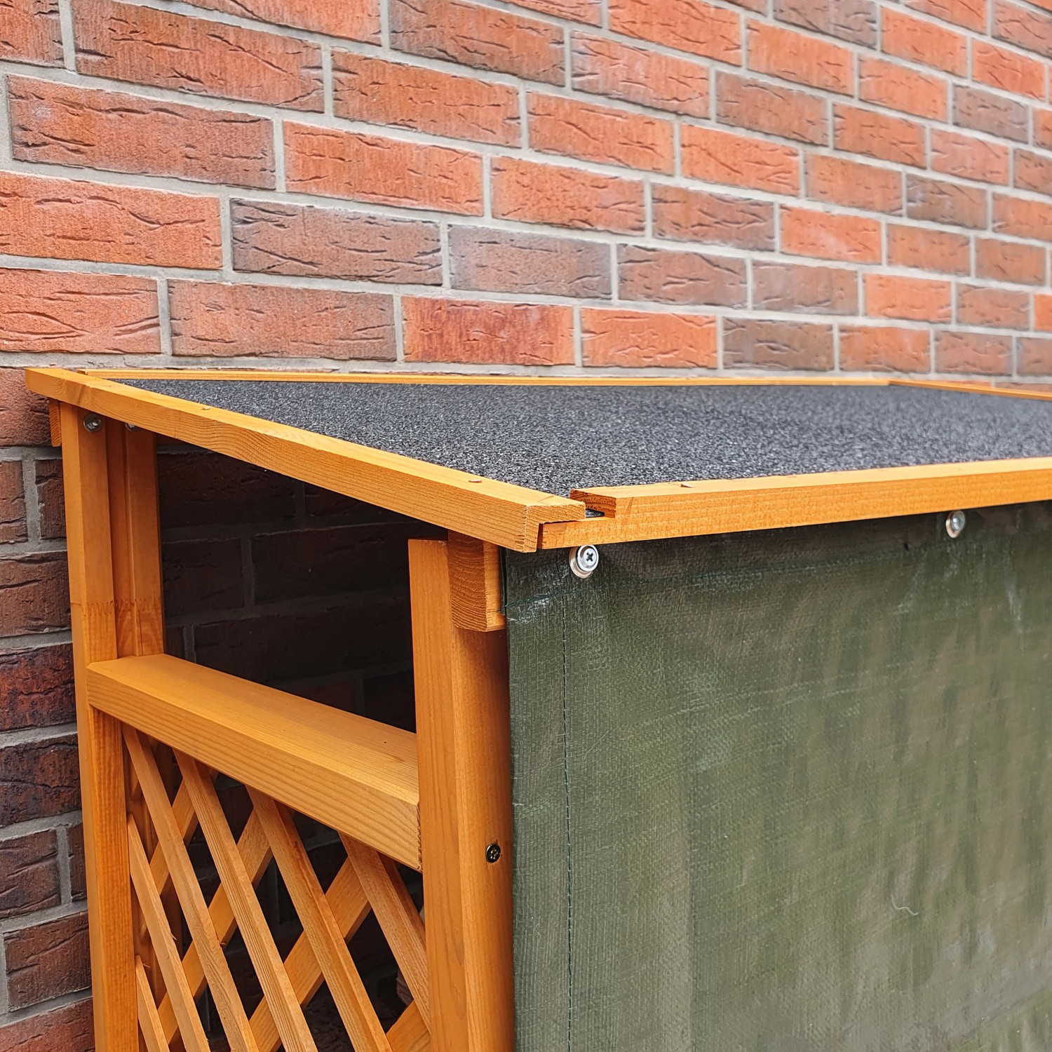 Kaminholzregal Speyer honigbraun mit Wetterschutzplane und Dachpappe vor einer roten Backsteinmauer.